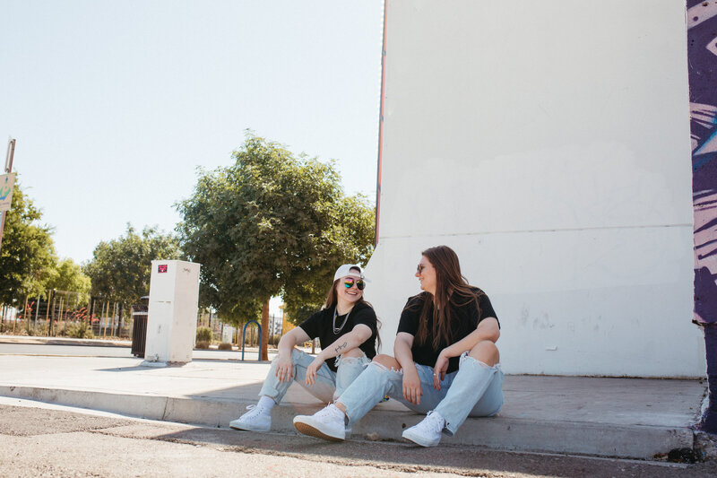 Two girls sit on a sidewalk, talking