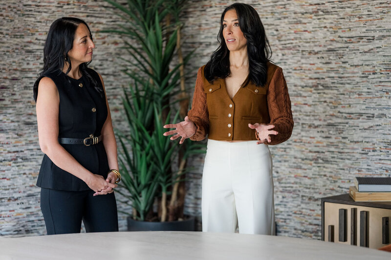 Two women standing, engaged in a conversation about sales techniques, with a plant nearby.