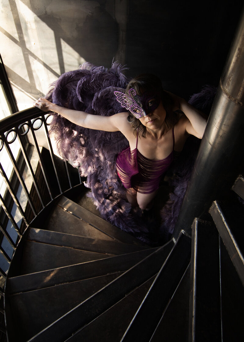 Woman in deep pink lingerie and feather robe posing confidently on a spiral staircase during a bold and artistic boudoir session with Christie Newell