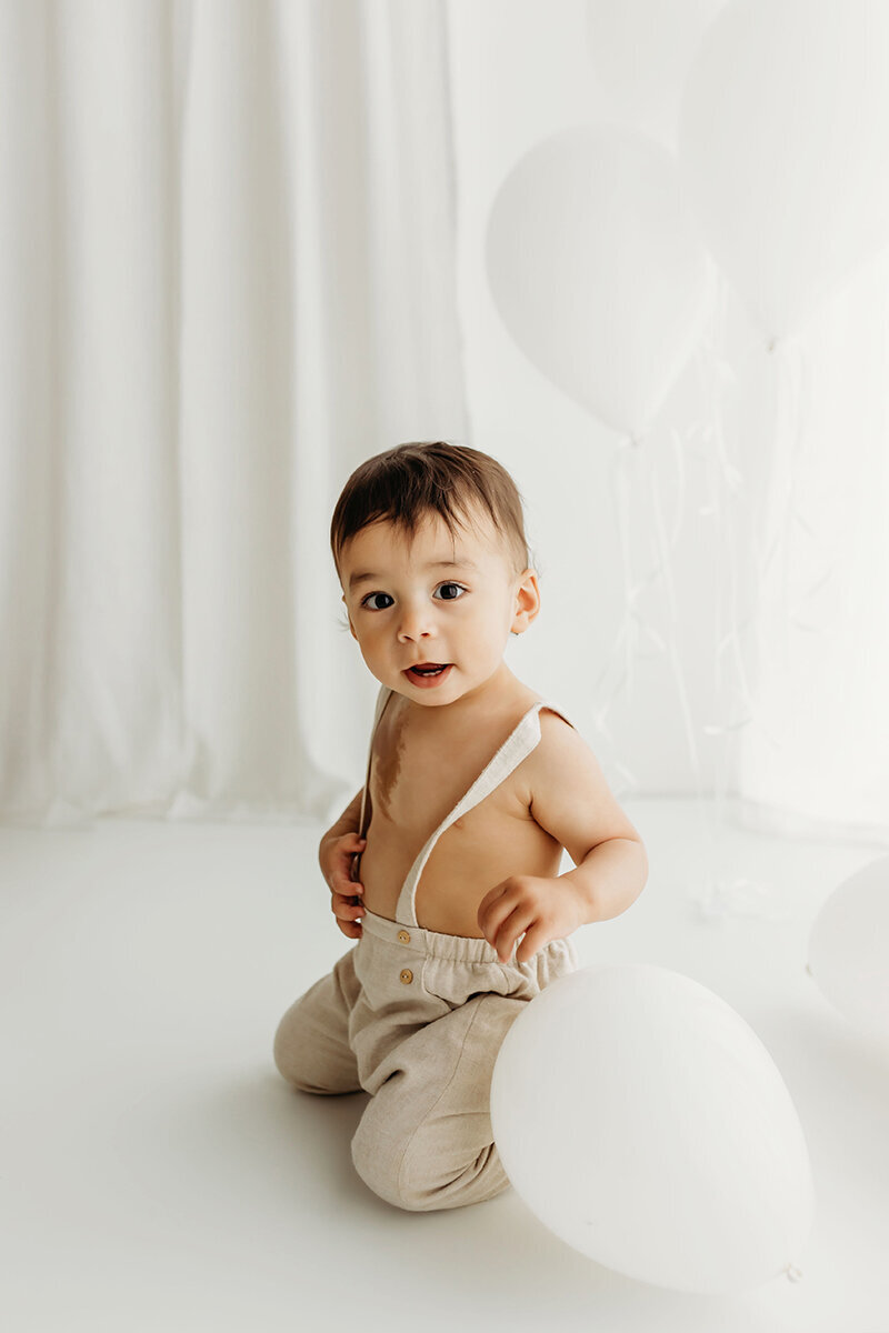 A year old boy smiles softly at the camera while playing with a white balloon during his first birthday photoshoot inside a natural light Vancouver Washington studio