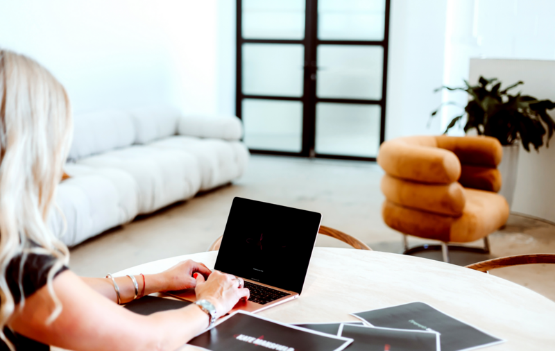 Woman working on a laptop at a round table in a modern, brightly lit office or studio space.
