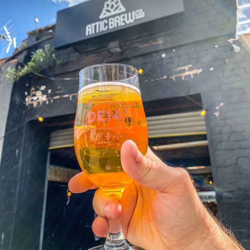 Hand holding a glass of craft beer up to a sign saying Attic Brew Co