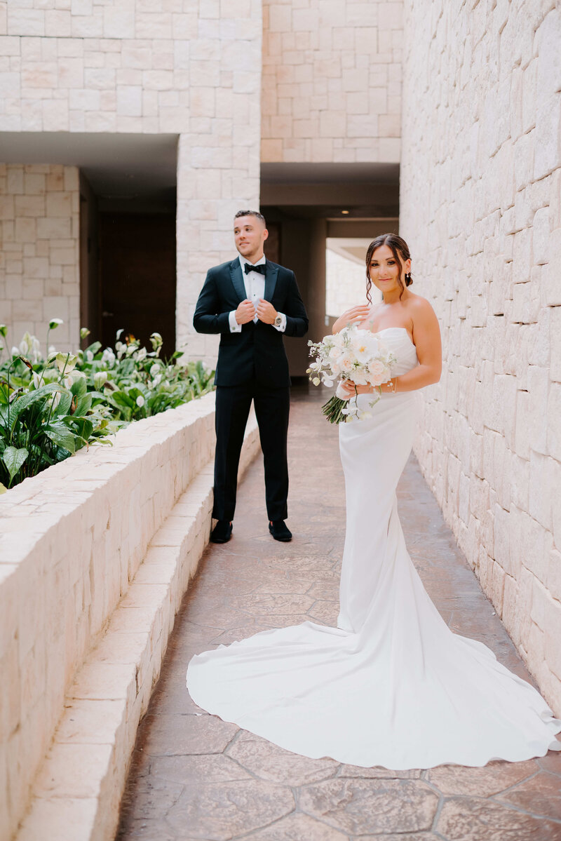 Elegant wedding couple portrait in a modern stone hallway, with the bride in a strapless white gown holding a bouquet and the groom in a black tuxedo adjusting his cufflinks