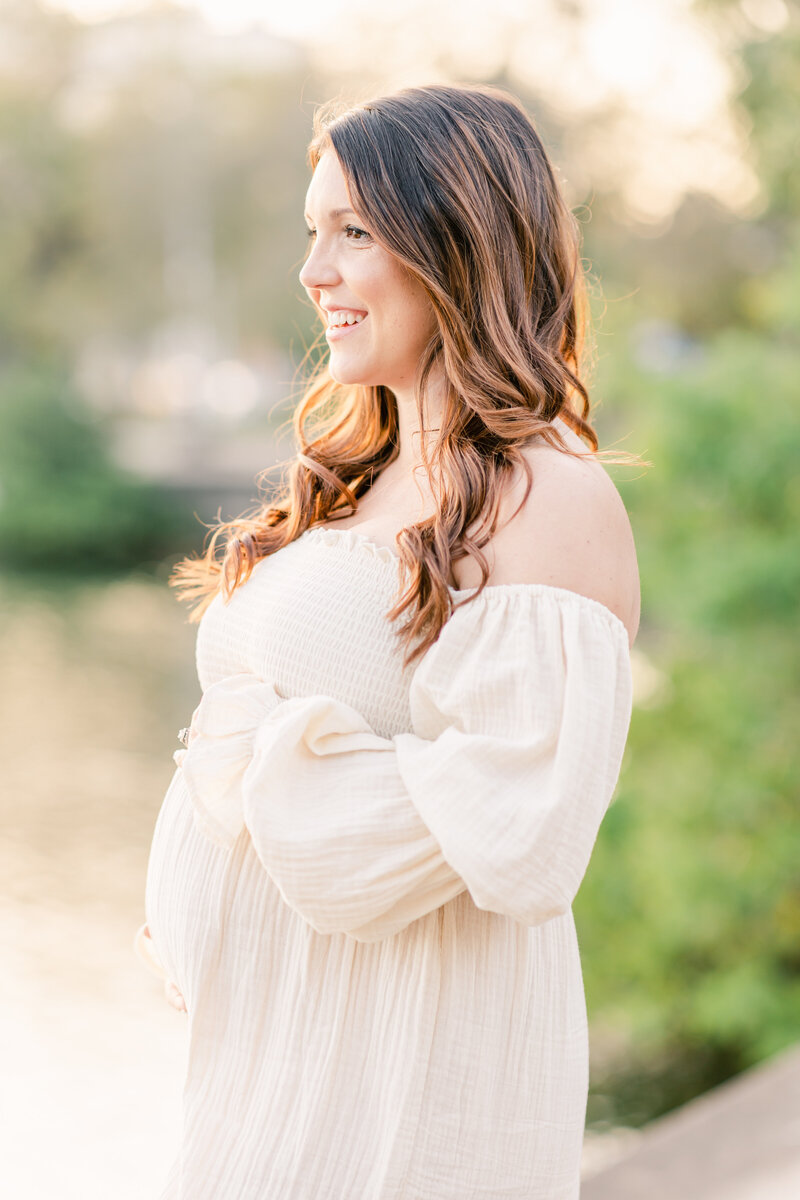 portrait of pregnant mother holding baby bump in white nothing fits but dress smiling off in the distance