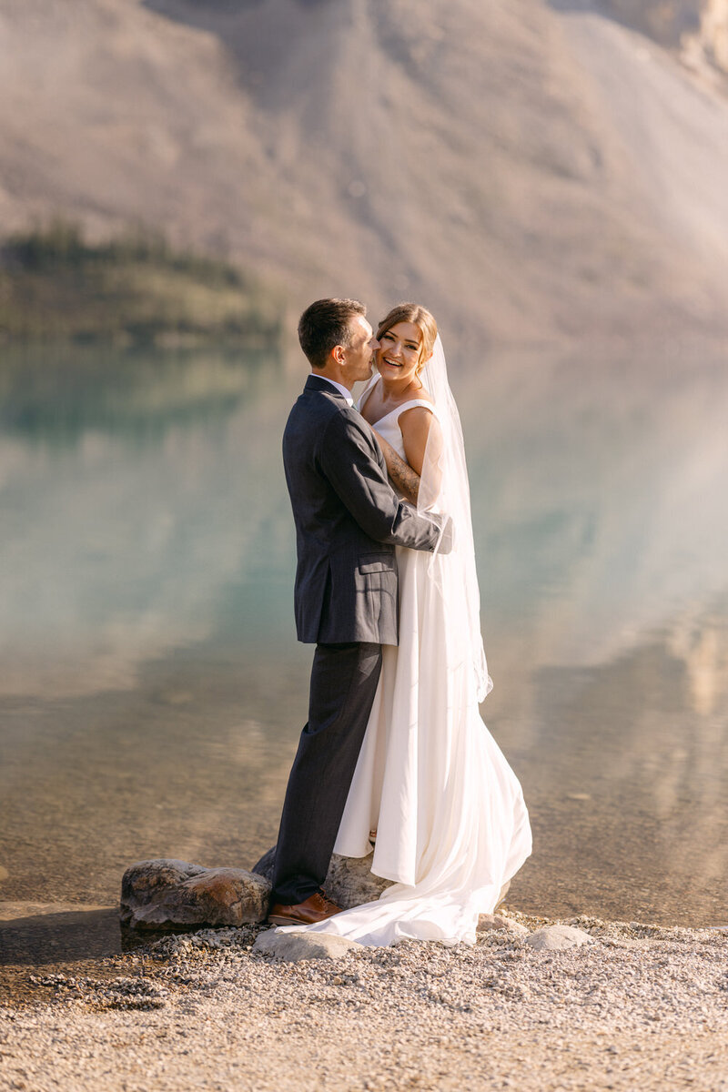 This image features a couple laughing joyously during their elopement beside Bow Lake in Banff National Park. The lush green setting and the tranquil backdrop of the lake enhance the intimate and joyful atmosphere of their special moment.