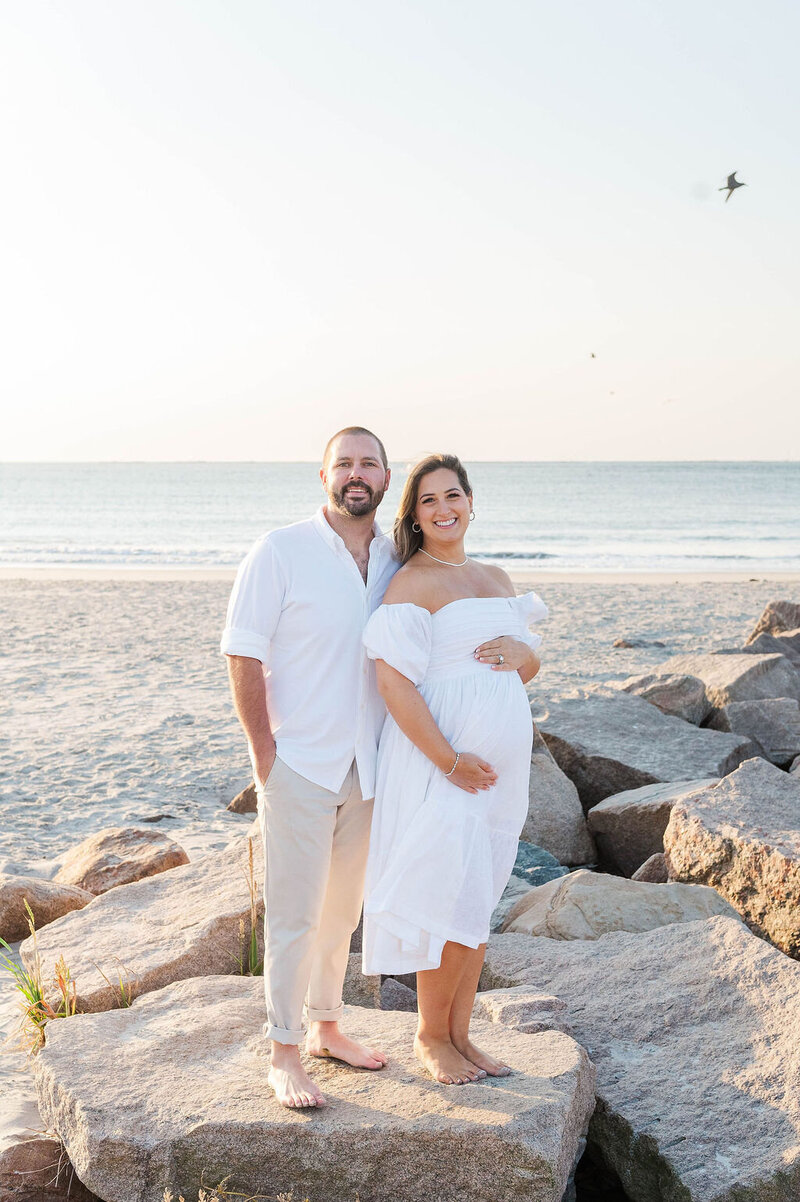 Pregnant woman holding her belly with husband behind her at beach in Narragansett, RI
