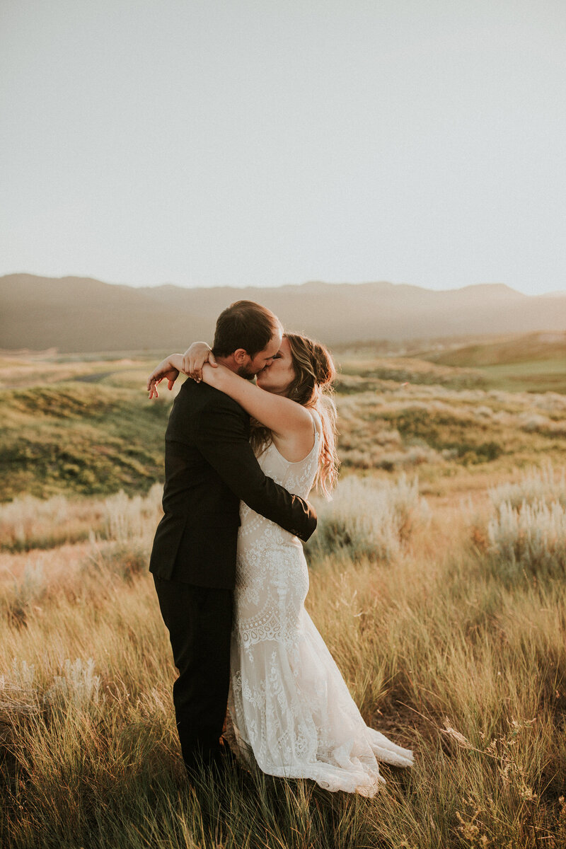 A bride and groom kiss while she wraps her arms around his neck in a golden field