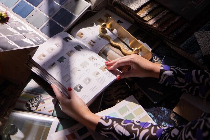 A person with red nail polish flips through a catalog of furniture designs, surrounded by swatches of fabric, paint, and other design materials.