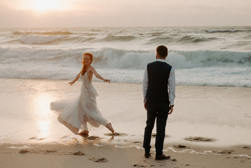Bride twirls on the beach while groom watches.