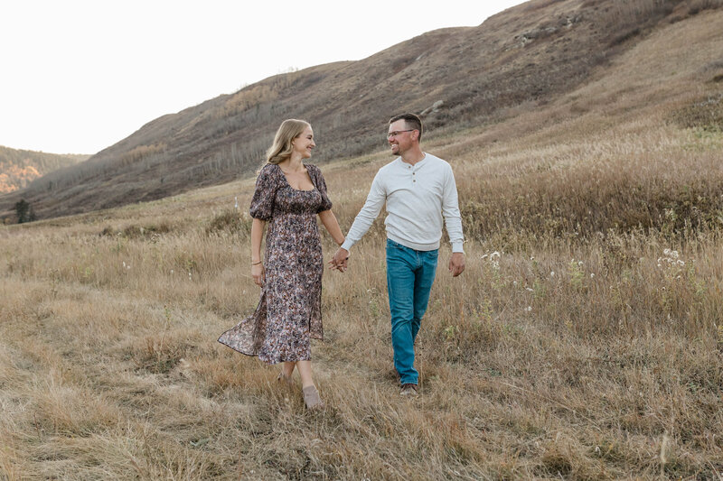 A young couple sit on driftwood on the shore of a mountain lake during their photography session. She has her hands clasped on one raised leg and is looking down and he's sitting beside her, kissing her on the side of her head.