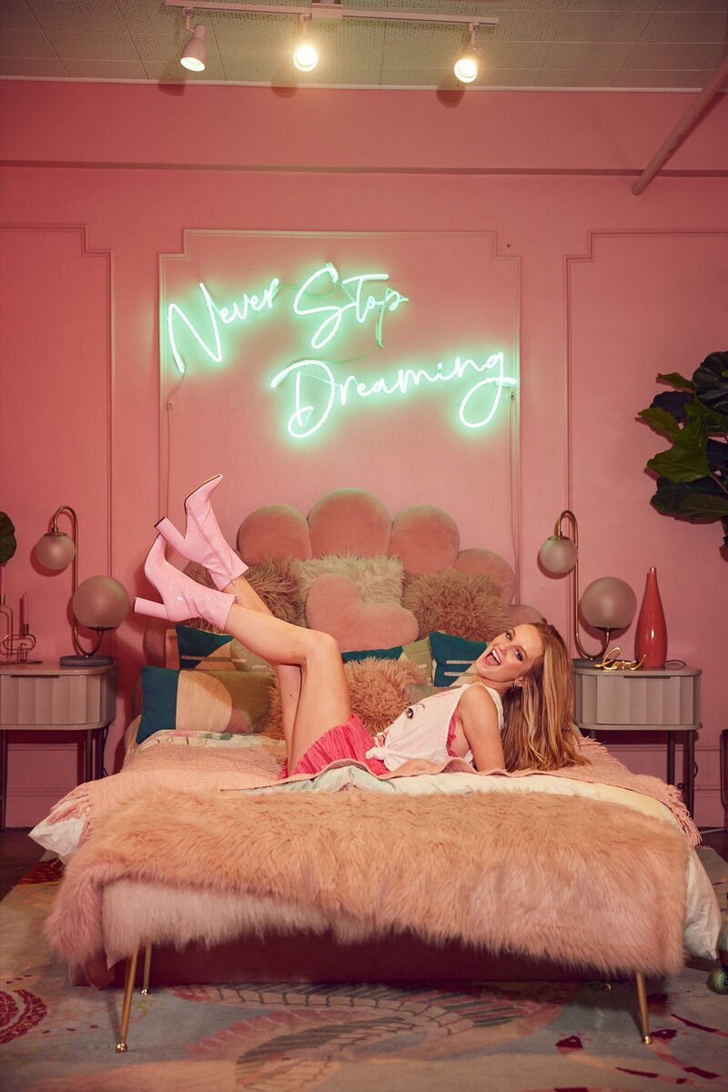 Playful and dreamy teen model lying on a fluffy bed under a “Never Stop Dreaming” neon sign in a colorful set – photographed during a styled workshop experience.