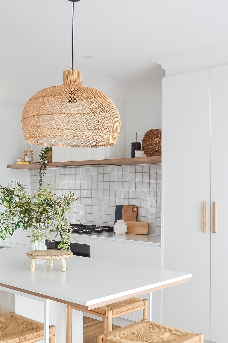 A woven rattan lamp gracefully illuminates the white table, adorned with a plant in a vase and a wooden stand, all nestled within the luxury home design ambiance crafted by a Newcastle interior designer.