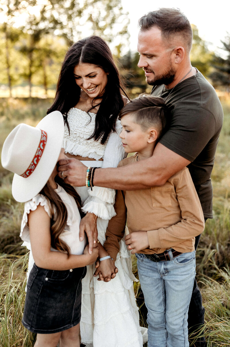 A beautiful family of 4 stands looking at each other as the father holds hi daughters face while smiling.  The family is in a scenic outdoor setting in Parker Colorado.