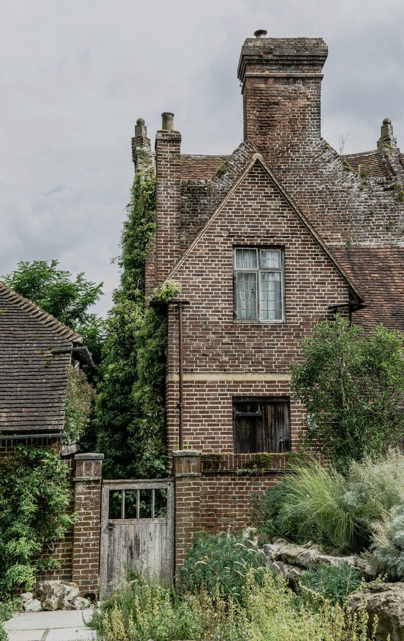 A historic brown brick house with aged, moss-covered bricks, partially hidden behind a matching brick fence and weathered timber gate. Green vines climb one side of the façade, while lush greenery and a natural rockery frame the foreground.