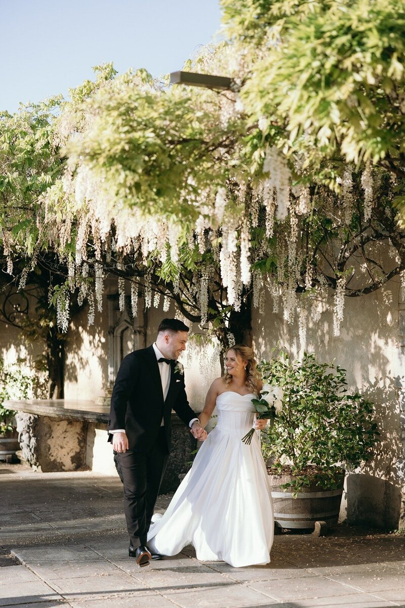 A bride and groom walking through an old courtyard with wisteria hanging down