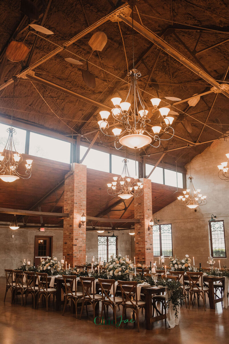 Industrial wedding venue with vaulted ceilings, brick columns, Crossback chairs, and long reception tables with white florals and taper candles.
