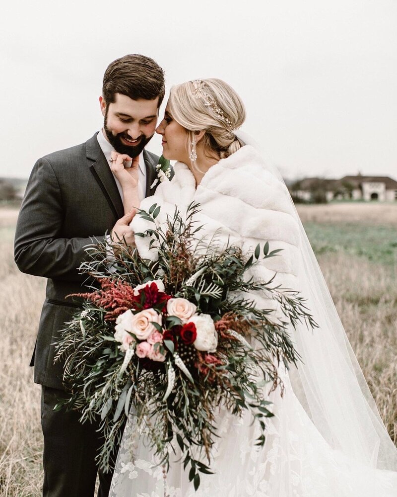 In a dimly lit hallway, the bride and groom sit on the floor amidst scattered petals, holding a bottle. Their graceful elegance is complemented by exquisite bridal hair and makeup packages, adding a touch of magic to their intimate moment.