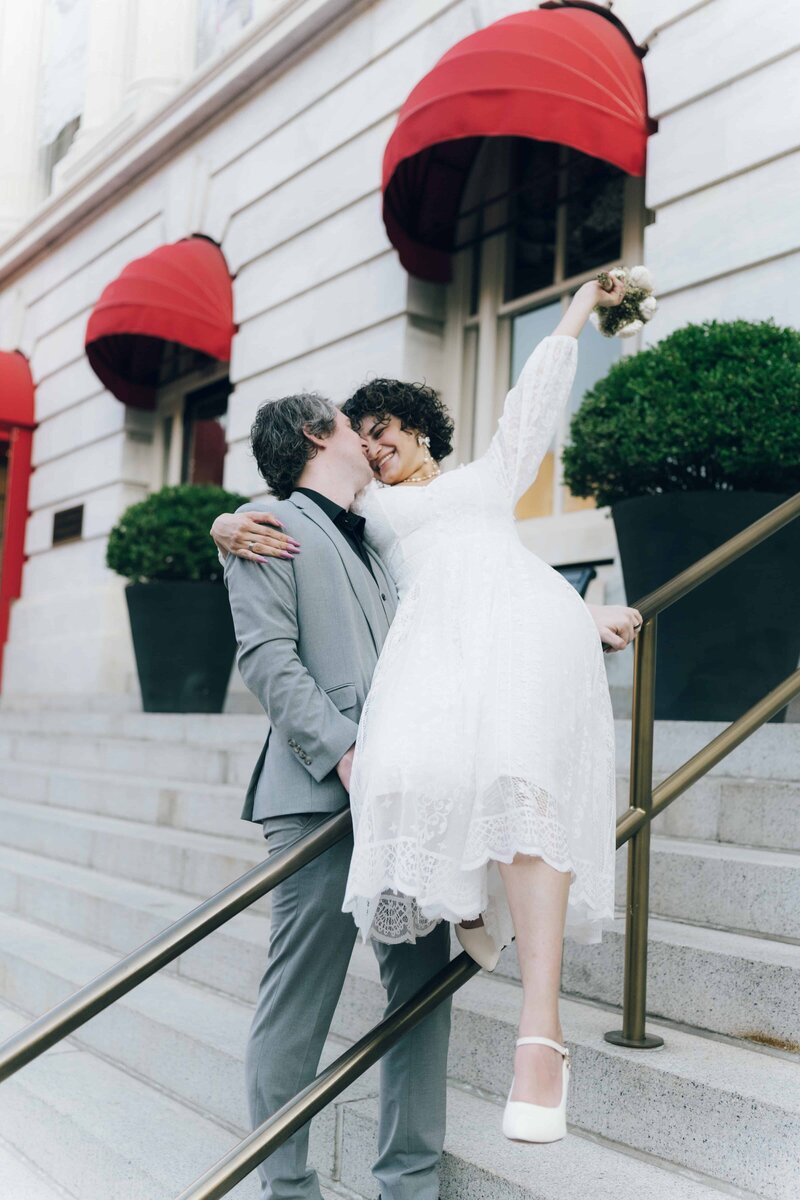 A bride in a white lace dress playfully leans over a railing, while her partner stands on the steps outside a building with red awnings.