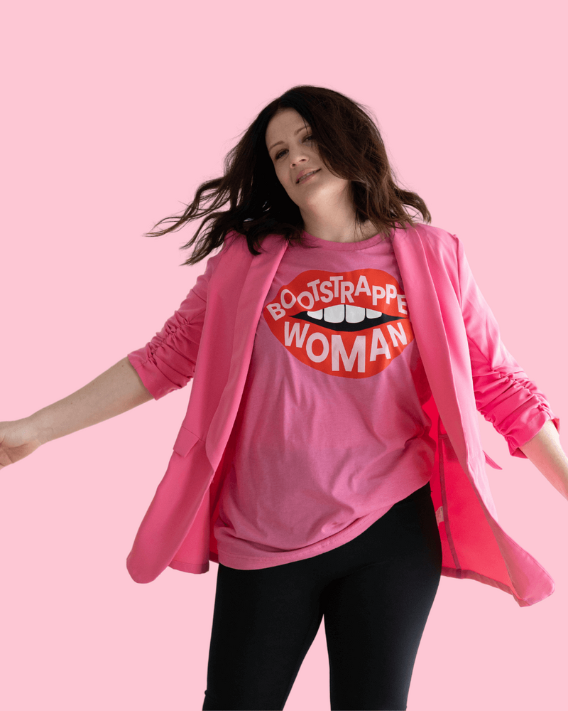 Emily, a brand and web designer, stands with arms outstretched in a pink shirt and jacket, radiating positivity.