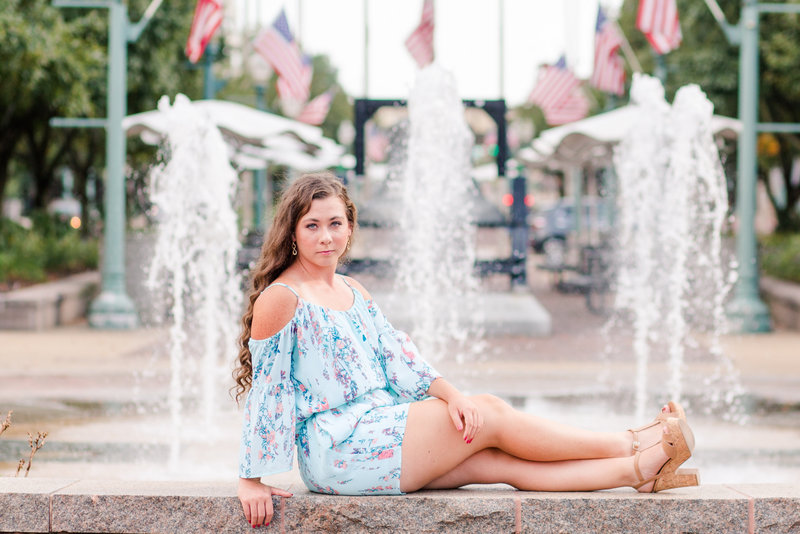 high school senior girl sitting on a ledge with fountains behind her, she has a cute light blue floral romper, with cute high heels, photographed by Jamie Lynette Photography Canton Ohio Senior Photographer