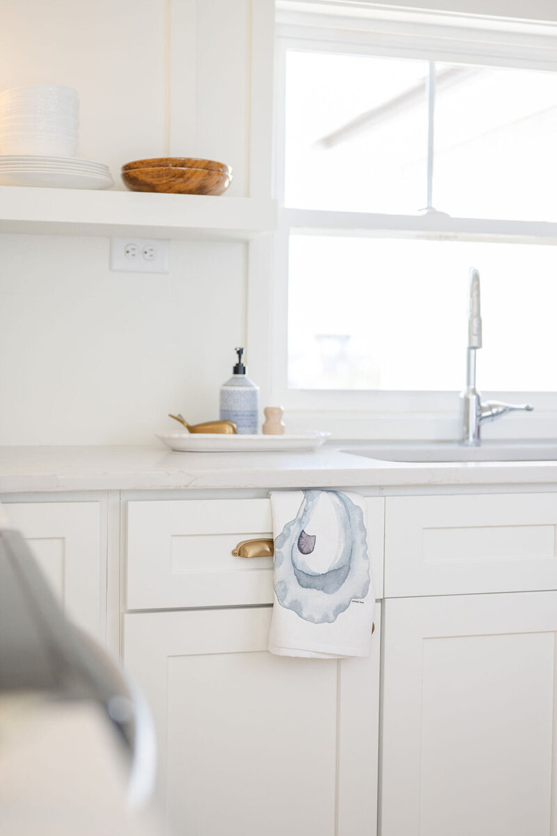 a pristine kitchen with white cabinets and shelves showcasing a watercolor painting of an oyster printed on a tea towel