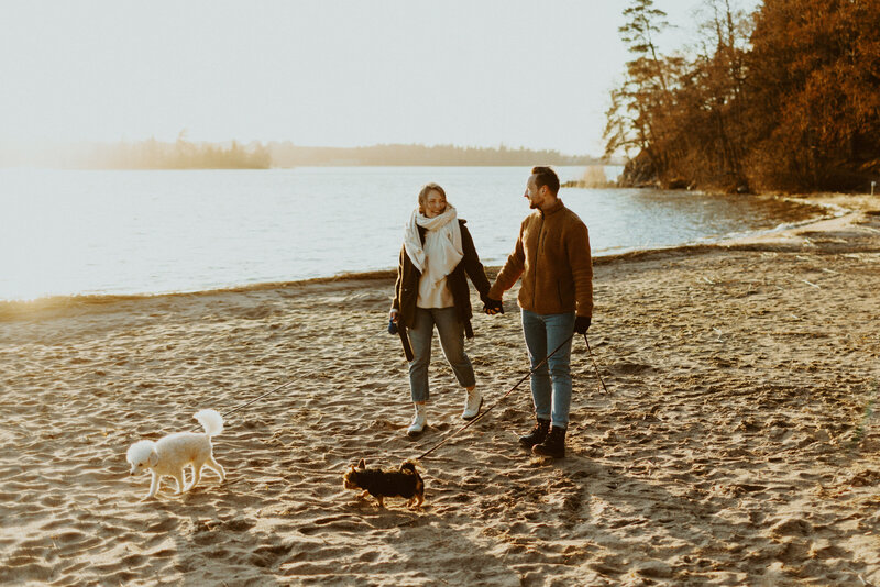 Engaged couple walking with their two little dogs at the beach looking at each other and smiling in Espoo in Finland.