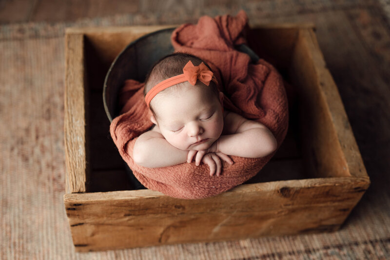 Newborn portraits in top newborn photographer studio. Baby girl is sleeping in a bucket draped with an apricot swaddle and matching headband. Baby's hands are folded under her chin and resting her head on her hands.