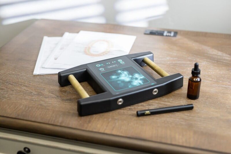 A Qest4 bioenergetic testing device with brass handholds on a desk beside a dropper bottle, reports, and a pen, used for energetic health assessments.