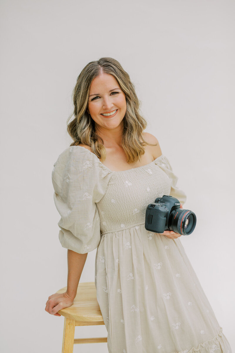 Lindsey Lambert brand photo, a smiling photographer in a soft neutral dress holding her camera in a natural light studio setting — Portrait photographer in Raleigh.