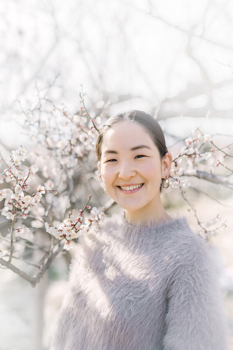 梅の花をバックに、明るく笑顔を見せている女性の写真。光が美しく降り注いでいて優しい印象。