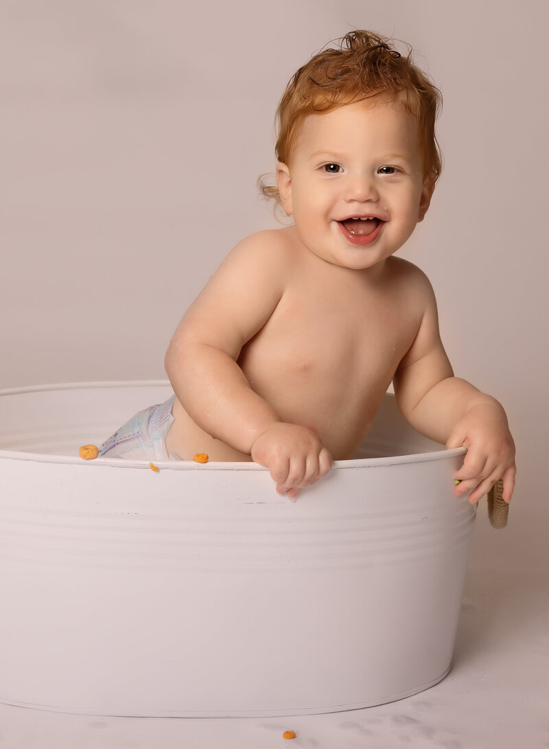 Baby boy smiles in a metal wash basin for a first birthday photoshoot. The baby has red curly hair and is smiling at the camera. Captured by premier Brooklyn NY family photographer Chaya Bornstein Photography.