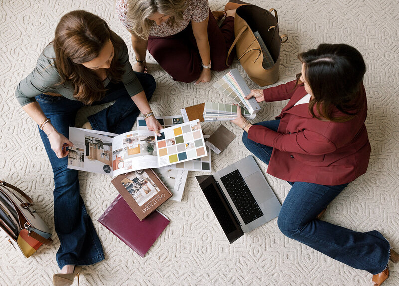 Three people are seated on the floor, looking at color swatches, paint samples, and design brochures. A laptop and a few bags are nearby. They appear to be discussing interior design choices on a patterned carpet.