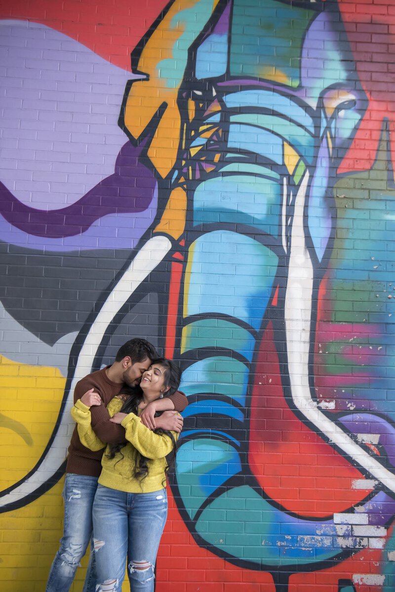 A couple hugging closely and standing in front of a mural on the side of a building 