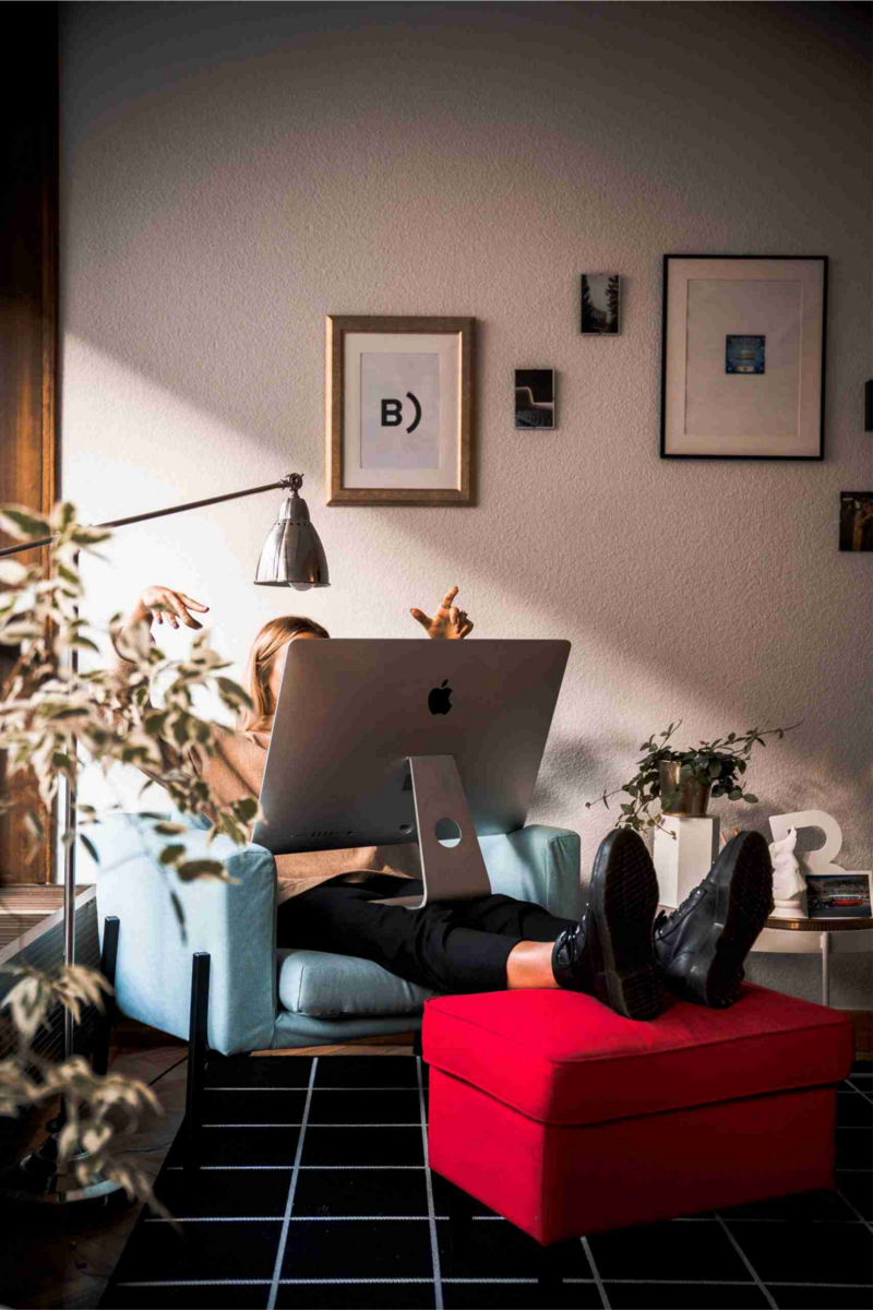 Shot of a woman in a decorated living space with color accents, hidden behind an iMac as she works, her feet up on a red ottoman.