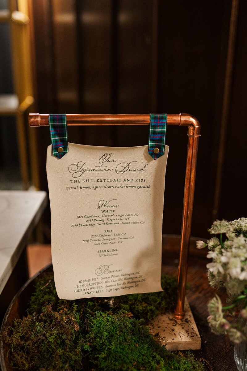 Fabric wedding bar menu hanging on a copper stand with tartan ribbon on a wine barrel next to the marble bar at District Winery in DC