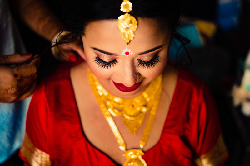 An indian bride looking down as her gold jewelry is applied by her friends before the wedding ceremony