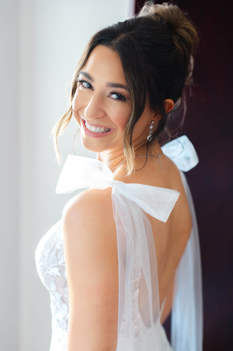 a bride in a white dress with bows on her shoulders looks over her shoulder smiling.  Captured by Ottawa wedding photographer JEMMAN Photography
