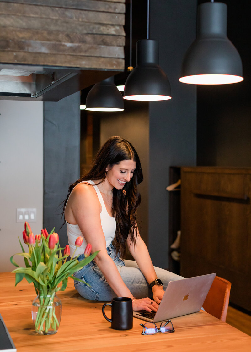 Allie Clark sitting at a kitchen counter with her laptop and coffee, smiling while working on a design project.