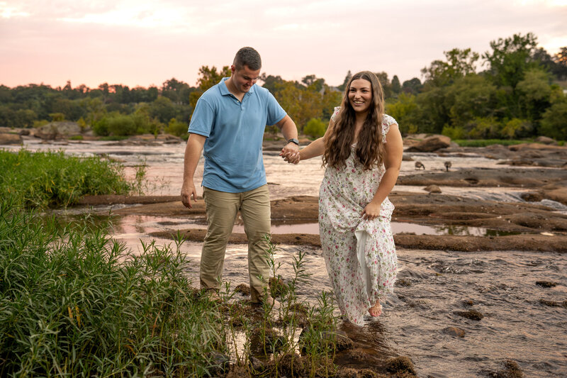 A women is leading her fiancé across rocks in the James river during their engagement shoot
