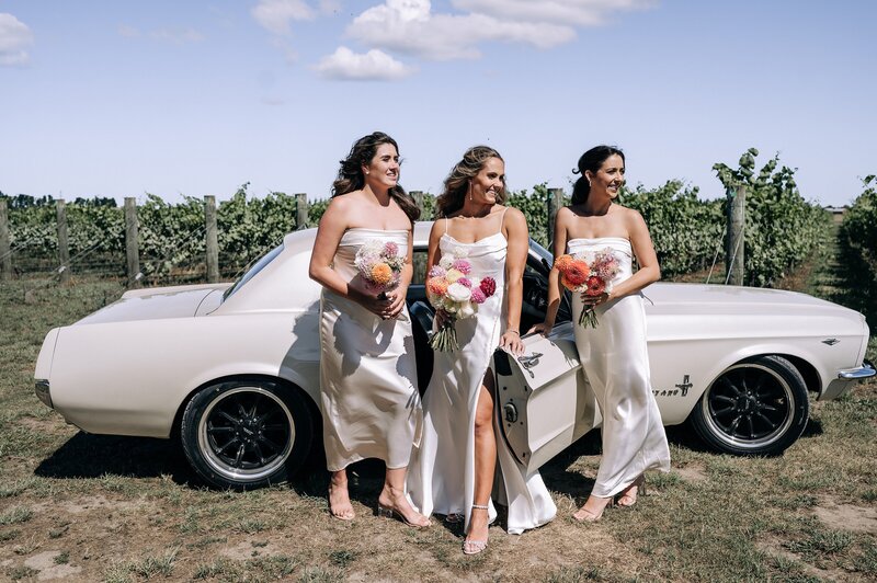 bride and bridesmaids in matching ivory with an ivory 1967 mustang on a vineyard in waipara christchurch nz