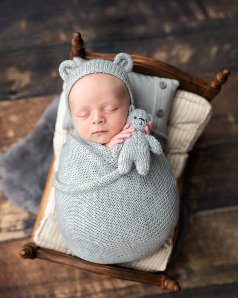 Soft blue knits and a tiny teddy—this newborn boy rests in pure comfort on a handcrafted baby bed.