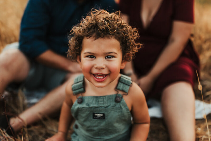 Curly headed boy in overalls smiles at the camera.