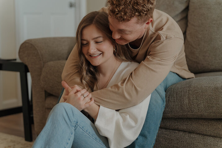 An intimate in home photographer session of a couple. The man is sitting on a couch with his girlfriend sitting between his legs and he holds her around her shoulders.