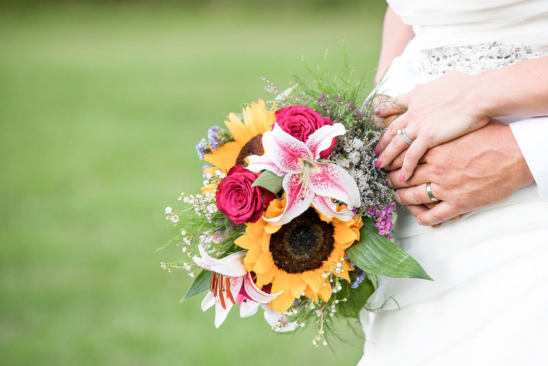 Close up of bride's and groom's hand with a bouquet featuring sunflowers, lilies, and ross