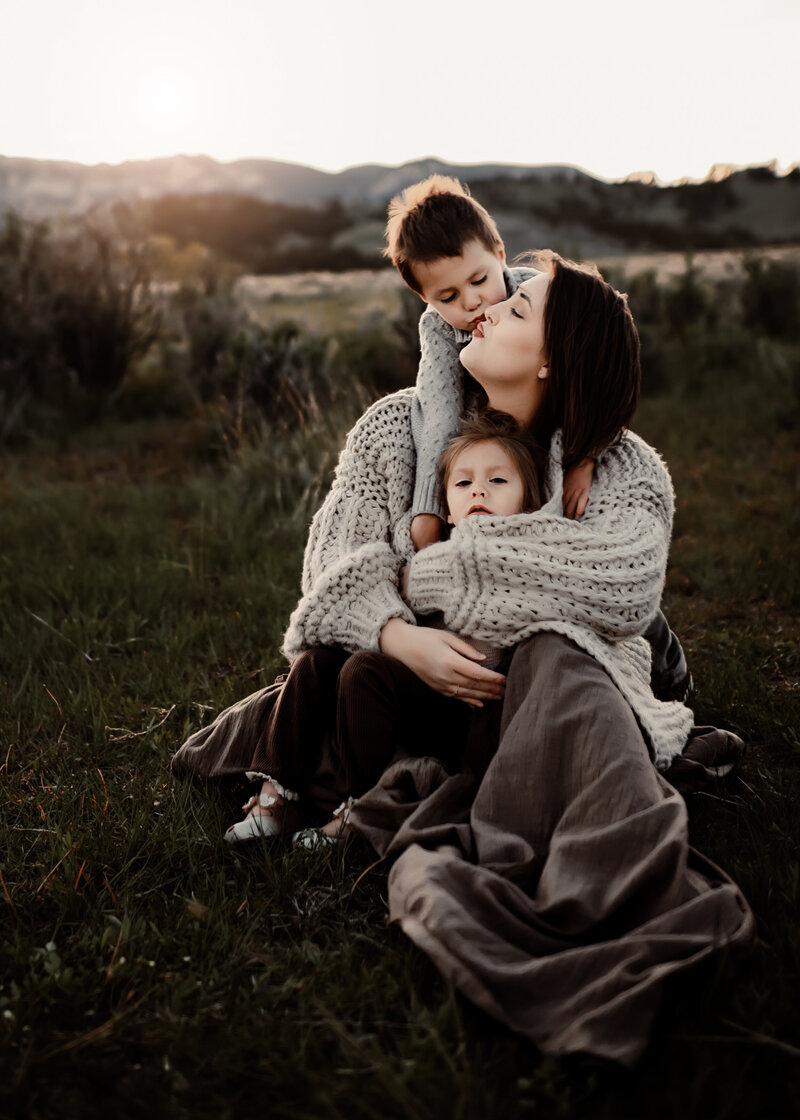 A mother holds her two young children in a lush green Wyoming field at sunset. 