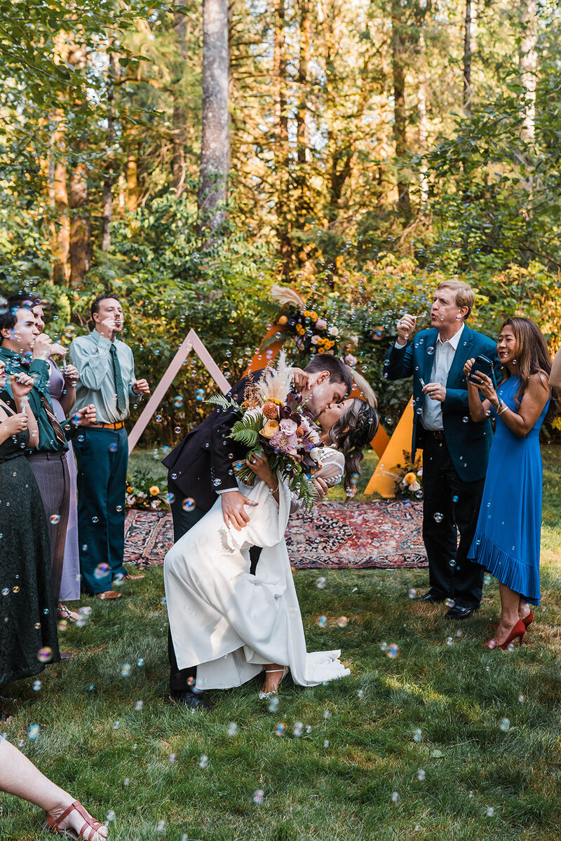 Groom dipping and kissing bride in front of colorful triangle ceremony arches while guests blow bubbles!