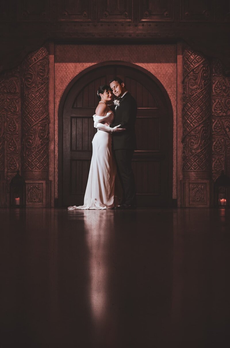 Bride and groom pose in front of the grand arched wooden doors at Madera Estates in Conroe, Texas, with rich red tones and intricate architectural details, captured in a cinematic wedding style by Logrono & Co.