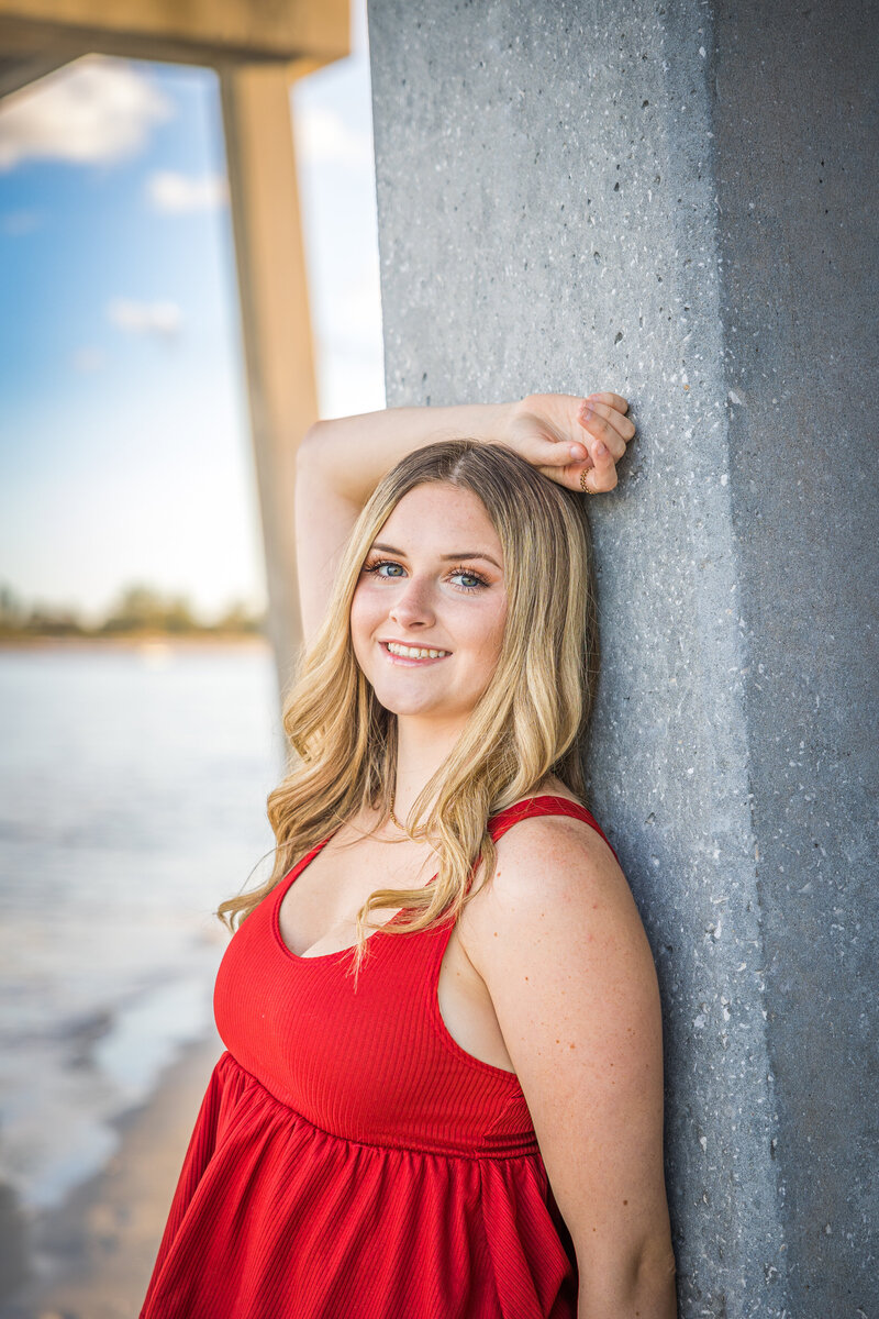 Senior posing under a bridge with her right arm up behind her head and wearing a red tank top.