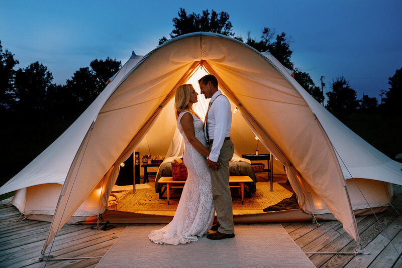 A bride and groom stand in front of their glamping tent at Bally Brook Farms. They are facing one another and holding hands. The Sun has just set, and the tent is glowing. Shenandoah Elopement photographer