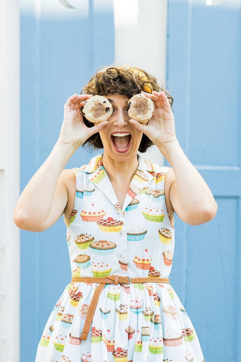 Playful female baker in a vintage-inspired dress covered in colorful pie illustrations holds two pastries over her eyes, laughing in front of a bright blue door. Her cheerful expression and whimsical outfit capture the fun and creativity of artisan baking. Small business branding photography in Columbia, SC, showcasing personality-driven branding for local bakers and food entrepreneurs.